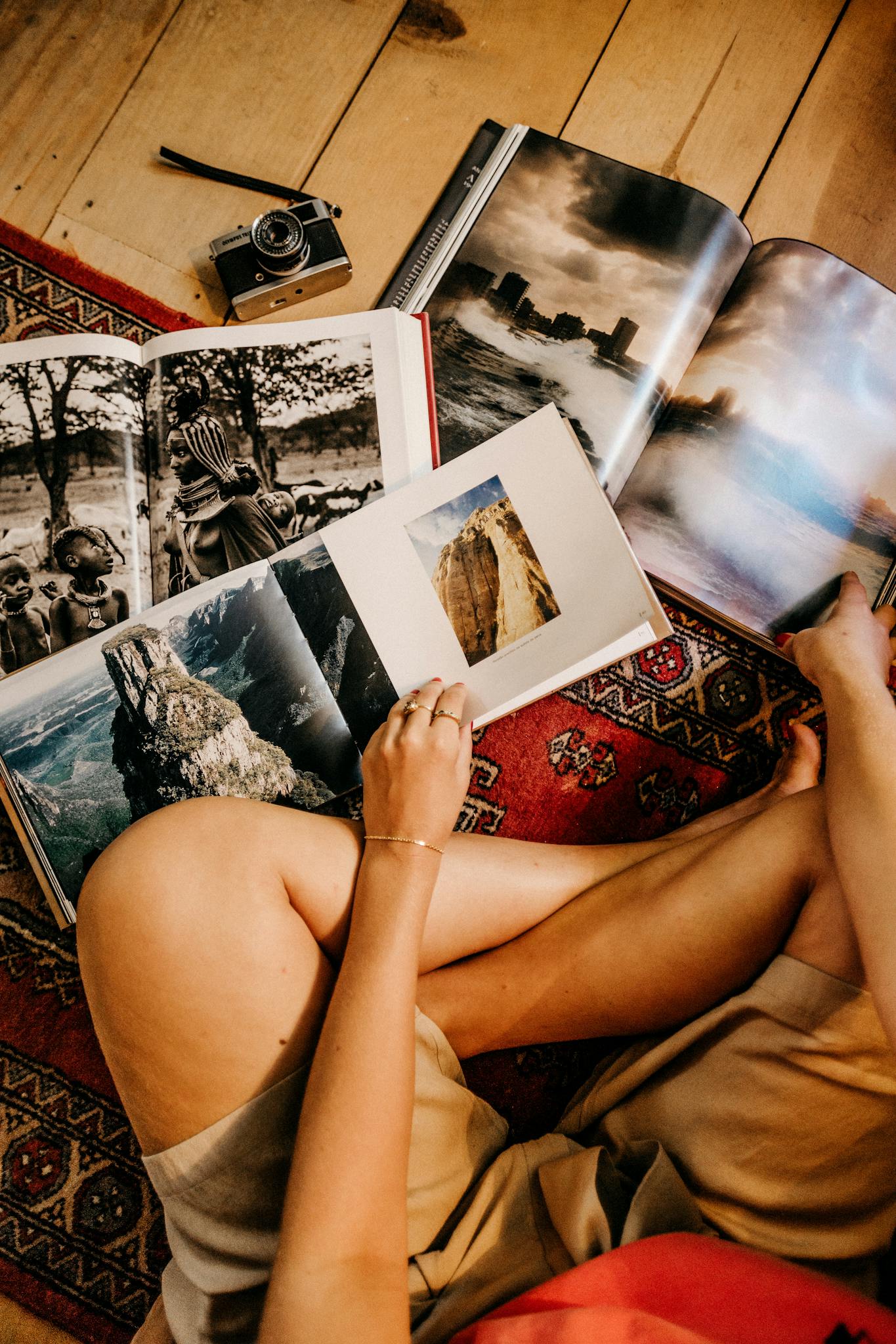 Person sitting on a rug, browsing photo books on a wooden floor with vintage camera nearby.